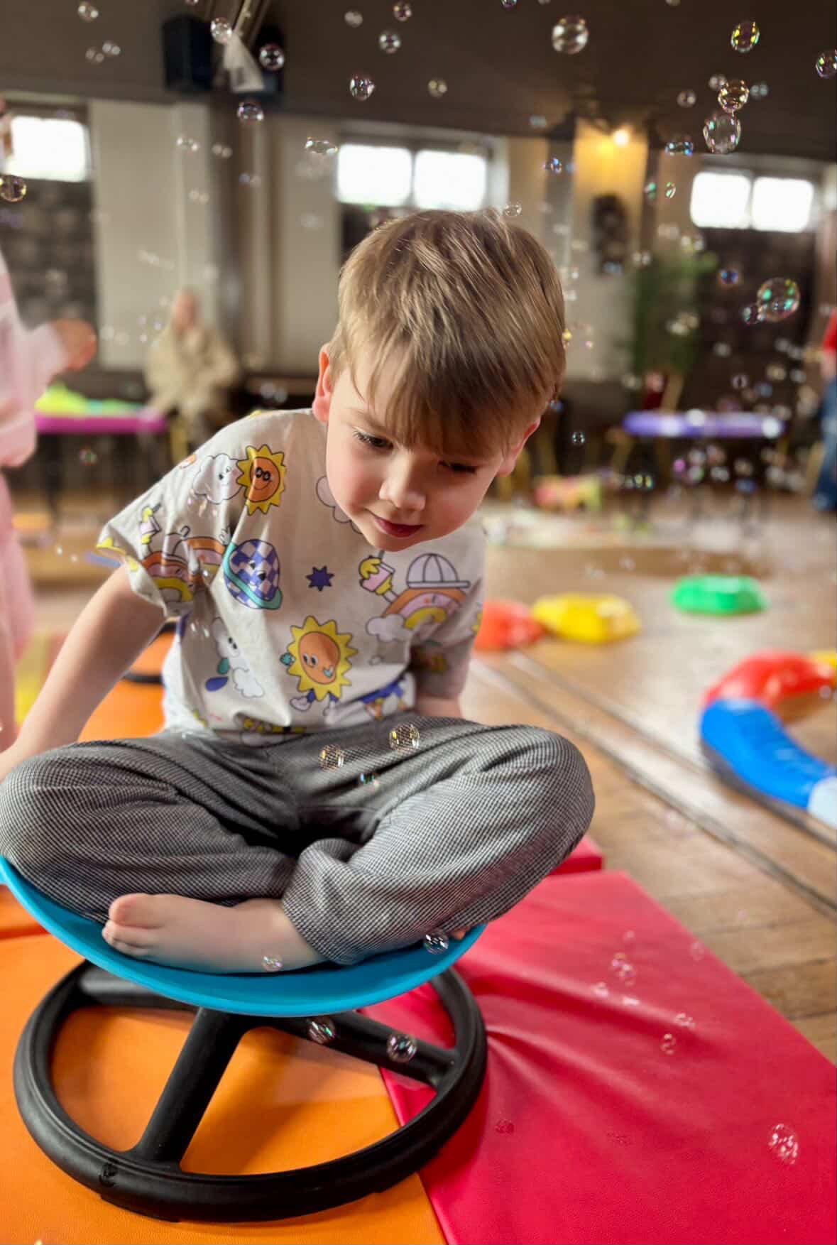A child attending a Fidget and Spin Stay and Play session sits on a spinning chair while watching floating bubbles.